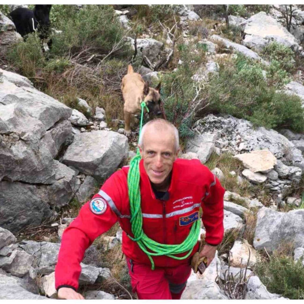 Un pompier en tenue rouge équipé d’un harnais de sécurité évolue sur une paroi rocheuse pour secourir deux chiens bloqués sur la montagne Sainte-Victoire.