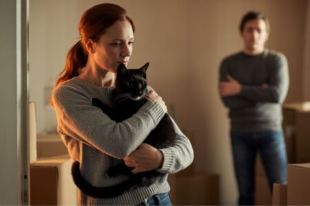 Juliette, une femme rousse, tient son chat noir Moka dans ses bras. En arrière-plan, un homme se tient bras croisés, l’air distant. Scène de séparation discrète.