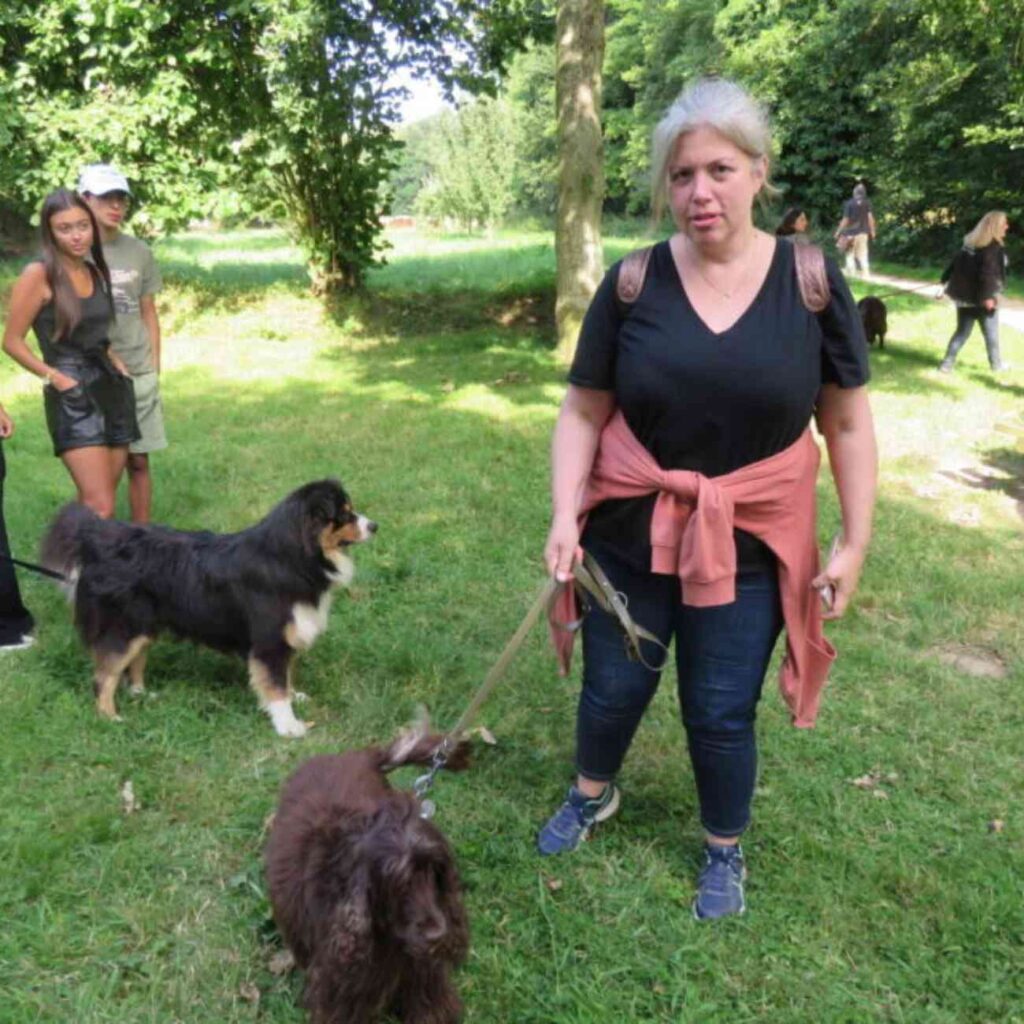 Femme tenant deux chiens en laisse dans un parc, entourée d’autres personnes et d’animaux, expression sérieuse
