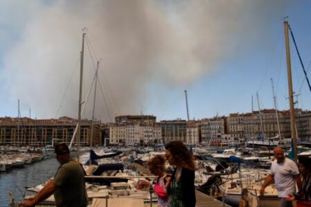Des habitants de Marseille rassemblés près d’un port, observant un épais nuage de fumée s’élevant au loin. L’ambiance est tendue. On distingue des chiens tenus en laisse au milieu de la foule. La scène se déroule dans un contexte d’incendie urbain.