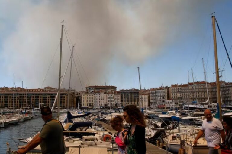 Des habitants de Marseille rassemblés près d’un port, observant un épais nuage de fumée s’élevant au loin. L’ambiance est tendue. On distingue des chiens tenus en laisse au milieu de la foule. La scène se déroule dans un contexte d’incendie urbain.