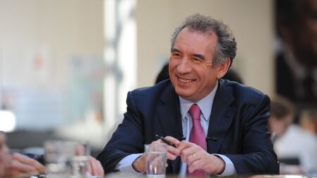 François Bayrou, en costume sombre, pose sérieusement devant un fond flouté lors d’un discours politique.