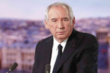 François Bayrou, en costume sombre, pose sérieusement devant un fond flouté lors d’un discours politique.