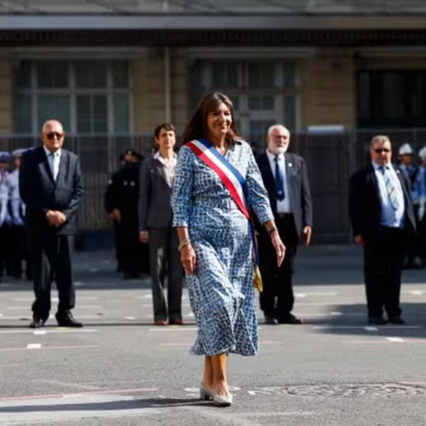 Anne Hidalgo marche en robe bleue dans la rue, entourée de plusieurs hommes en costume, lors d’un déplacement officiel