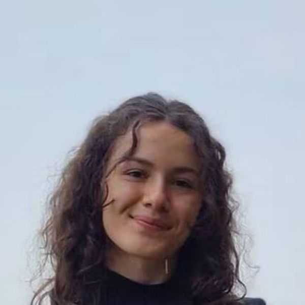 Portrait de Camille Cagnot, doctorante en psychologie et éthologie animale à l’Université de Caen Normandie, photographiée en extérieur sur fond de ciel bleu clair. Elle sourit doucement, les cheveux longs et bouclés, vêtue d’un haut noir.