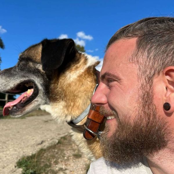 Portrait d’un homme souriant aux côtés d’un chien, tous deux regardant dans la même direction en plein air.