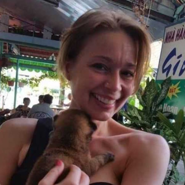 Personne souriante tenant un petit animal dans ses bras, en extérieur sous un auvent végétalisé.